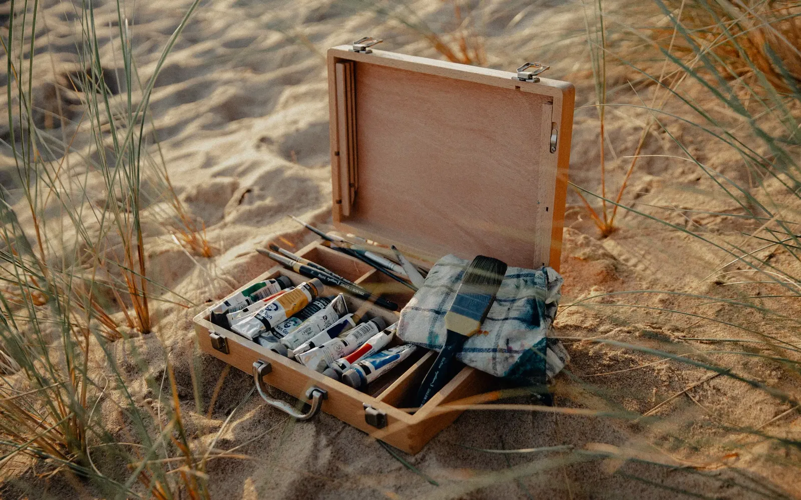 A wooden suitcase of paints, laying on sand at a beach.