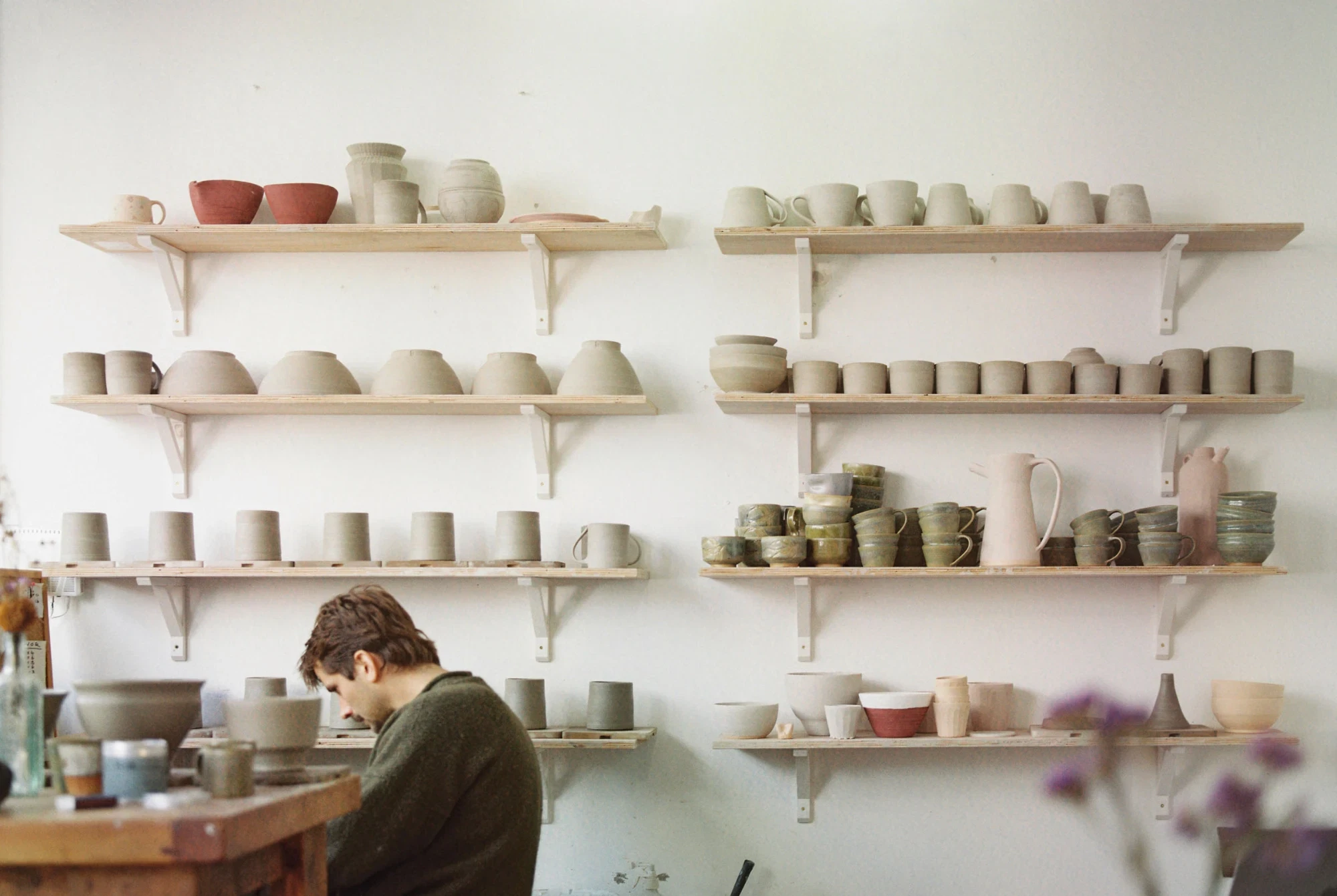 Louis sat a potters wheel with shevles and his pots drying in the background.